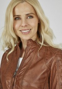 Braune Kunstlederjacke mit hohem Kragen, sichtbarem Reißverschluss und strukturierten Details entlang der Nähte. Präsentiert sich mit glatter, glänzender Oberfläche.