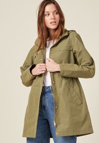 Giacca impermeabile verde oliva con cappuccio, zip frontale, tasche laterali e bottoni a pressione. Caratterizzata da una vestibilità rilassata e una superficie liscia.