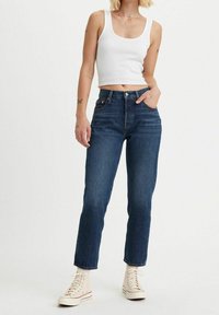 Jeans de denim azul de tiro alto con diseño de pierna recta, que cuentan con bolsillos frontales y cierre de botón. Combinados con una camiseta sin mangas de canalé blanca.