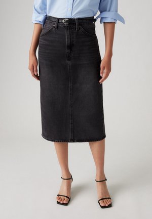 Femme portant une chemise bleu clair rentrée dans une jupe midi en jean noire avec une ceinture noire et des sandales à talons noires.
