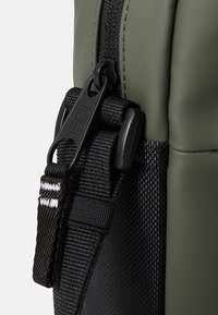 Olivenfarbene Tasche aus mattem Material, mit einem schwarzen Gurt und einem Plastikreißverschluss mit strukturierten Details und reflektierenden Akzenten.