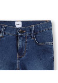 Jeans de denim azul escuro com um design clássico de cinco bolsos, apresentando um botão preto e costura laranja. Etiqueta lê "BOSS."
