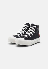 Czarne wysokie trampki z płótna, z białymi sznurowadłami, białymi gumowymi podeszwami oraz czerwonymi sercami i gwiazdami na zewnętrznych bokach.