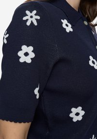 Tmavě modré polo tričko s krátkými rukávy a bílými květinovými vzory, vyrobené z strukturované pleteniny s vlnitým lemem na spodním okraji. Kšiltd dvěma knoflíky.