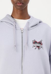 Sudadera con cremallera de color gris claro que presenta un gráfico frontal con un corazón rojo, el texto "Converse" y elementos florales; textura suave con capucha y cordones.