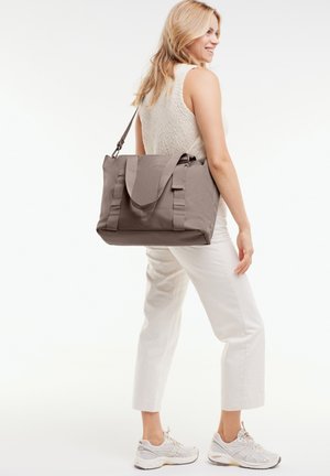 Femme portant un haut sans manches en maille et un pantalon blanc, portant un grand sac fourre-tout gris sur l'épaule, souriante et regardant sur le côté.