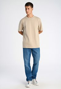 Beige t-paita lyhythihaisena yhdistettynä sinisiin farkkuihin. Asu on rento ja siihen kuuluu valkoiset tenniskenkä.