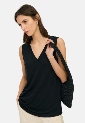 Mujer con cabello largo y castaño que lleva una blusa negra sin mangas con escote en V y pantalones claros a rayas, sosteniendo un bolso de hombro negro tejido.