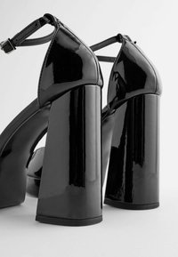 Chaussures à talons hauts en cuir verni noir avec un talon épais, un bout pointu et une bride à la cheville, présentant une finition lisse et brillante avec peu de détails.
