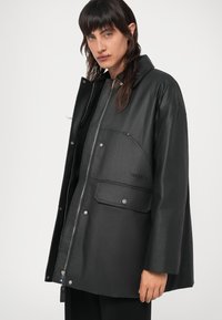 Casaco preto, oversized, feito de um material sintético liso. Apresenta um colarinho alto, botões de pressão prateados, um bolso de remendo e detalhe do logo.