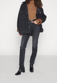 Chaqueta de mezclilla negra superpuesta a un suéter de cuello alto a rayas marrón y naranja, combinado con jeans oscuros y botas de tobillo negras y brillantes.