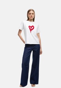 Witte katoenen t-shirt met een rood hartontwerp met druipaccenten, gecombineerd met donkere jeans met rechte pijpen. Model staat tegen een lichte achtergrond.