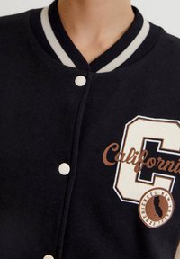 Černá univerzitní bunda s kontrastním šedým a bílým žebrovaným límcem, patentkami a logem "California" hnědé a bílé barvy na hrudi.