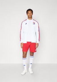 Weiße Sportjacke mit roten und blauen Streifen, rote Shorts, weiße Socken mit blauen Streifen und weiße Fußballschuhe. Enthält das Teamlogo auf der Jacke.