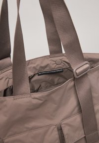 Sac fourre-tout marron fabriqué à partir de filets de pêche recyclés, doté de poignées robustes, d'un compartiment zippé et de poches à patch à l'avant pour le rangement.