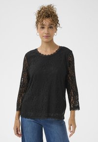 Sort blondelags top med bølget halsudskæring og trekvartærmer. Mønstrede blondeoverflader over en solid bund, båret med blå jeans.