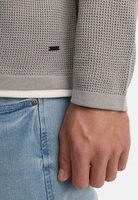 Suéter de punto gris con un patrón texturizado, presenta puños de canalé. Llevado sobre una camisa blanca, combinado con jeans de mezclilla azul claro.