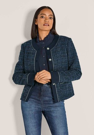 Vrouw met steil haar in een marineblauwe textuurjas over een gerimpelde blouse met hoge kraag en blauwe jeans, staande tegen een effen achtergrond.