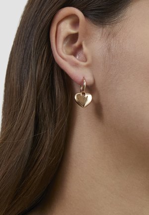 Nahaufnahme eines Ohrs einer Frau mit glattem braunem Haar, die einen goldenen Creolenohrring mit einem hängenden herzförmigen Anhänger trägt.