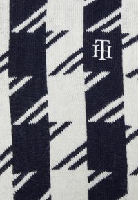 Stickad tröja med ett vitt och marinblått zigzag-mönster, med en liten logotyp i vitt. Mjuk textur och medelhållfasthet.