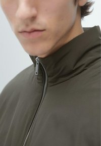 Giacca verde oliva con colletto alto e cerniera argentata, caratterizzata da una texture liscia ed elegante, design aderente e dettagli minimalisti.