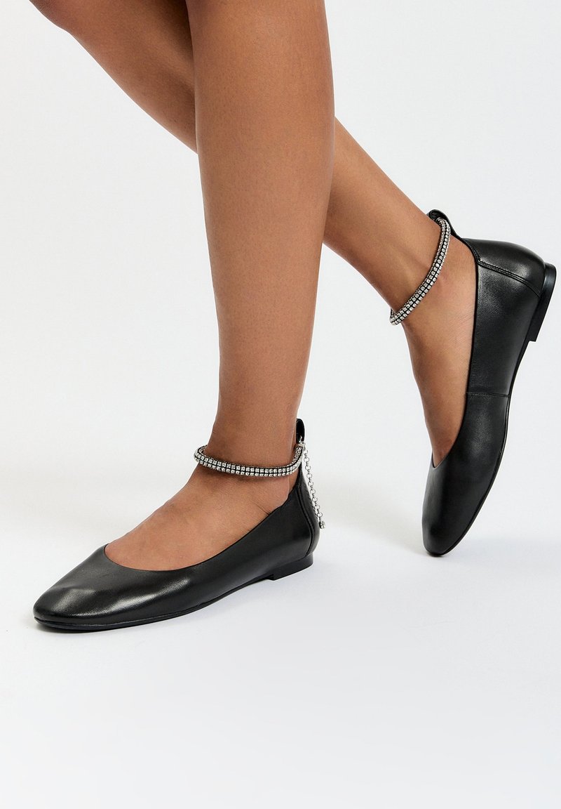 Bailarinas de piel negra con punta redonda, que presentan una correa en el tobillo adornada con detalles de cadena en tono plateado. Textura suave y diseño minimalista.