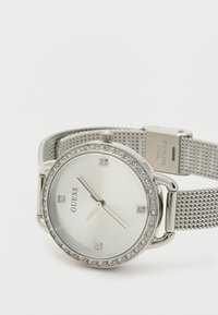 Relógio com bracelete em malha prata, mostrador redondo branco, realces de cristal em torno do mostrador e marcadores de horas prateados. Logótipo da marca "GUESS" exibido.