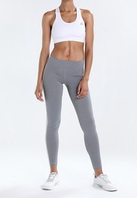 Žena v bílém sportovním podprsenkovém topu, šedých legínách s vysokým pasem a bočními pruhy, a bílých sportovních teniskách, stojící na neutrálním pozadí.
