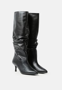 Botine negre din piele, cu înălțime până la genunchi, cu vârfuri ascuțite, cu gambe căzute și tocuri mici, prezentate una lângă alta pe un fundal alb.