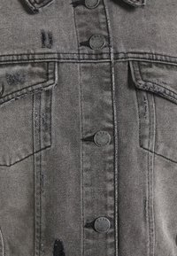 Šedá džínsová bunda s odřenými detaily, čtyřmi kapsami a černými knoflíky. Má klasický límeček a viditelné švy.