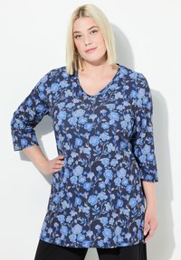 Marineblaue Blusentunika mit 3/4-Ärmeln, versehen mit hellblauen Blüten und grünen Akzenten, aus weichem, dehnbarem Stoff. V-Ausschnitt-Design.