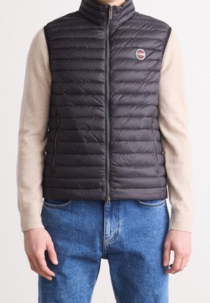 Mies, jolla on musta tikattu vetoketjullinen liivi beigejen pitkähihasten ja sinisten farkkujen päällä, näyttää etuviiston, kädet rentoina sivuilla.