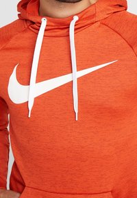 Homme portant un sweat à capuche Nike orange avec des cordons blancs et un grand logo Nike swoosh blanc sur la poitrine.