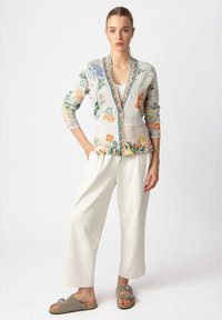 Cardigan lavorato a maglia con motivi floreali in verde, arancione e giallo, dotato di scollatura a V e bordi a contrasto, abbinato a pantaloni beige larghi e sandali.