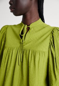 Blusa verde, strutturata, con scollo rotondo, tessuto plissettato e un dettaglio con lacci sul petto. Le maniche a sbuffo aggiungono volume. Materiale leggero.