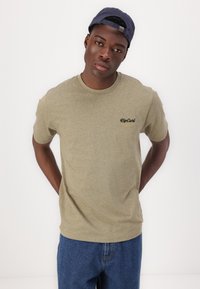 T-shirt in cotone verde oliva con maniche corte e colletto rotondo, caratterizzato da un logo nero "RipCurl" sul lato sinistro. Abbinato a jeans blu.