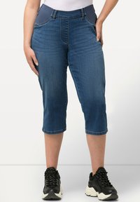 Pantalones capri de mezclilla en azul oscuro con una amplia cinturilla elástica, bolsillos frontales y dobladillos terminados. Usados con zapatillas negras.