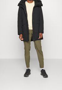 Parka larga negra con capucha, cierre de cremallera y bolsillos frontales; combinada con camisa beige clara y pantalones verde oliva, rematada con zapatillas negras.