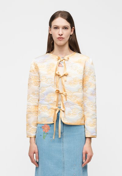 Femme portant une veste matelassée jaune pâle avec des motifs de nuages et des liens en ruban, associée à une jupe en jean bleu avec une broderie florale.