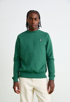Green sweatshirt with a crew neck and ribbed cuffs, featuring a small logo on the chest. Worn with light striped pants.