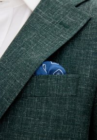 Vihreä teksturoitu bleiseri, jossa on viilto-olkain ja taskuliina, joka on sinisestä kankaasta valkoisella kukkakuviolla.