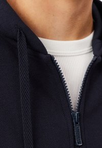 Marineblauwe hoodie met een gestructureerde stof, volledige ritssluiting en capuchon. Gedragen over een wit geribd shirt, met een zichtbaar ritstrekkerdetail van het merk.