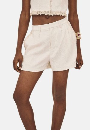 Pantalones cortos blancos de flores, hechos de una tela texturizada, con una cintura ajustada, pliegues frontales y bolsillos laterales. Un detalle de encaje adorna el dobladillo.