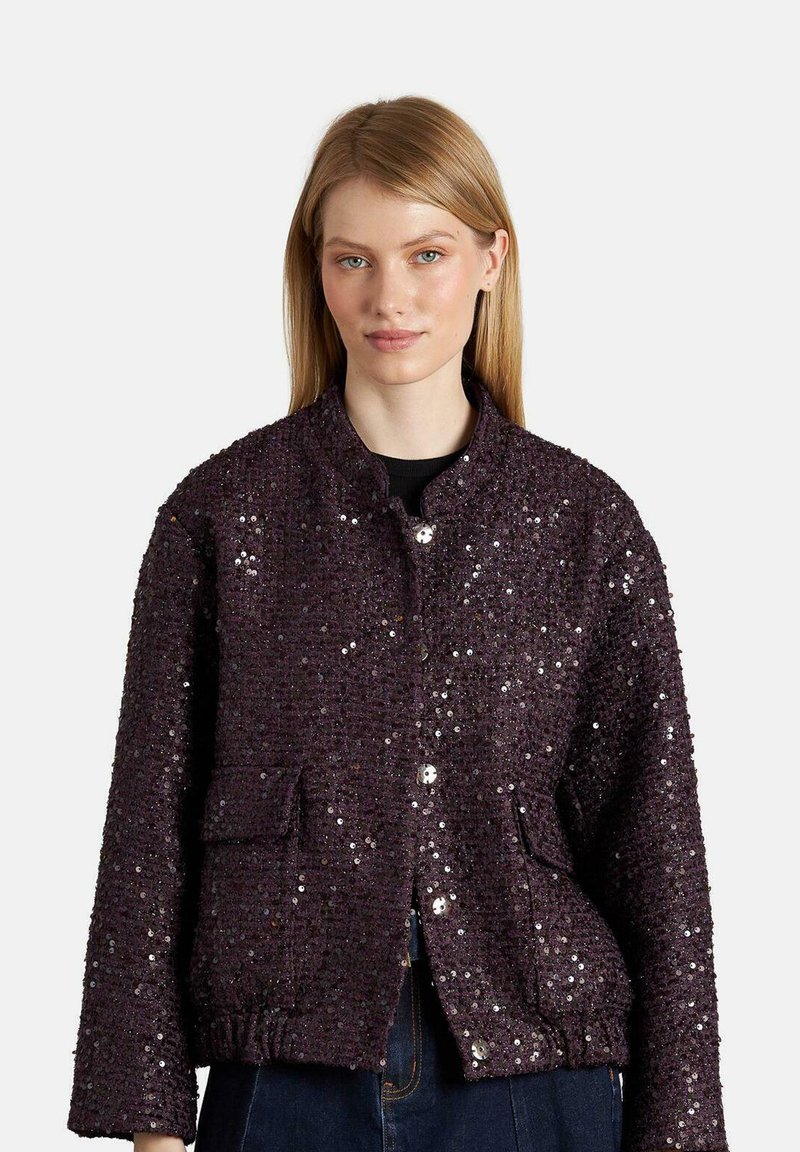 Giovane donna che indossa una giacca di paillettes viola scuro con chiusura a bottoni, tasche a soffietto e orlo elastico, in piedi davanti a uno sfondo bianco.