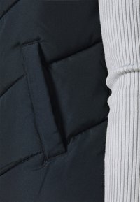 Czarna pikowana kamizelka z boczną kieszenią; częściowo widoczny na prawej stronie jasnoszary, prążkowany rękaw swetra. Miękka, gładka faktura materiału.