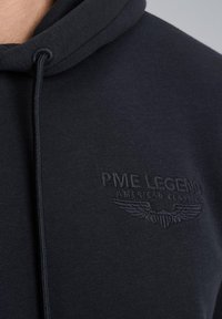 Hoodie van donkere marineblauwe stof; voorzien van een getextureerde borduursel van "PME LEGEND" en het vleugellogo op de borst. Koordjes in de capuchon.