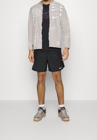 Chaqueta gris claro con cremallera y diseños gráficos, camiseta negra, pantalones cortos negros y zapatillas grises con detalles en naranja.