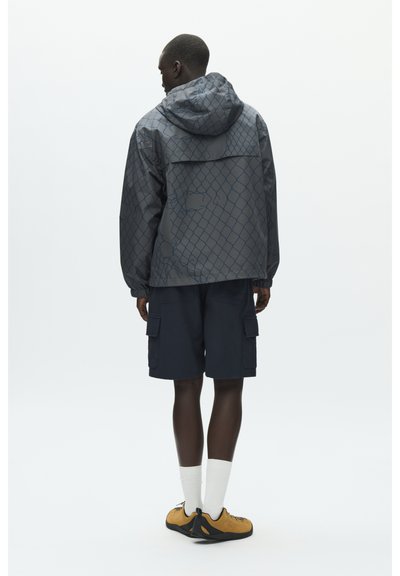 Homme portant une veste à capuche grise à motifs, un short cargo bleu marine, des chaussettes blanches et des baskets jaunes et noires, debout de dos.
