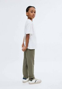 Ung kille som står på sidan med händerna knäppta bakom ryggen, iklädd en vit t-shirt med tryck, olivgröna byxor och vita sneakers med blå ränder.
