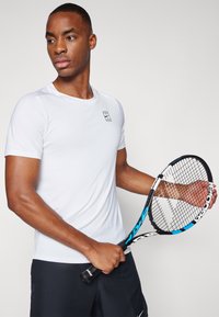 White short-sleeve athletic shirt with a black logo on the chest, worn with dark shorts; holding a tennis racket with a blue accent.